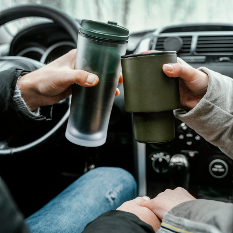 En la imagen se observan dos manos sujetando termos de café dentro de un auto