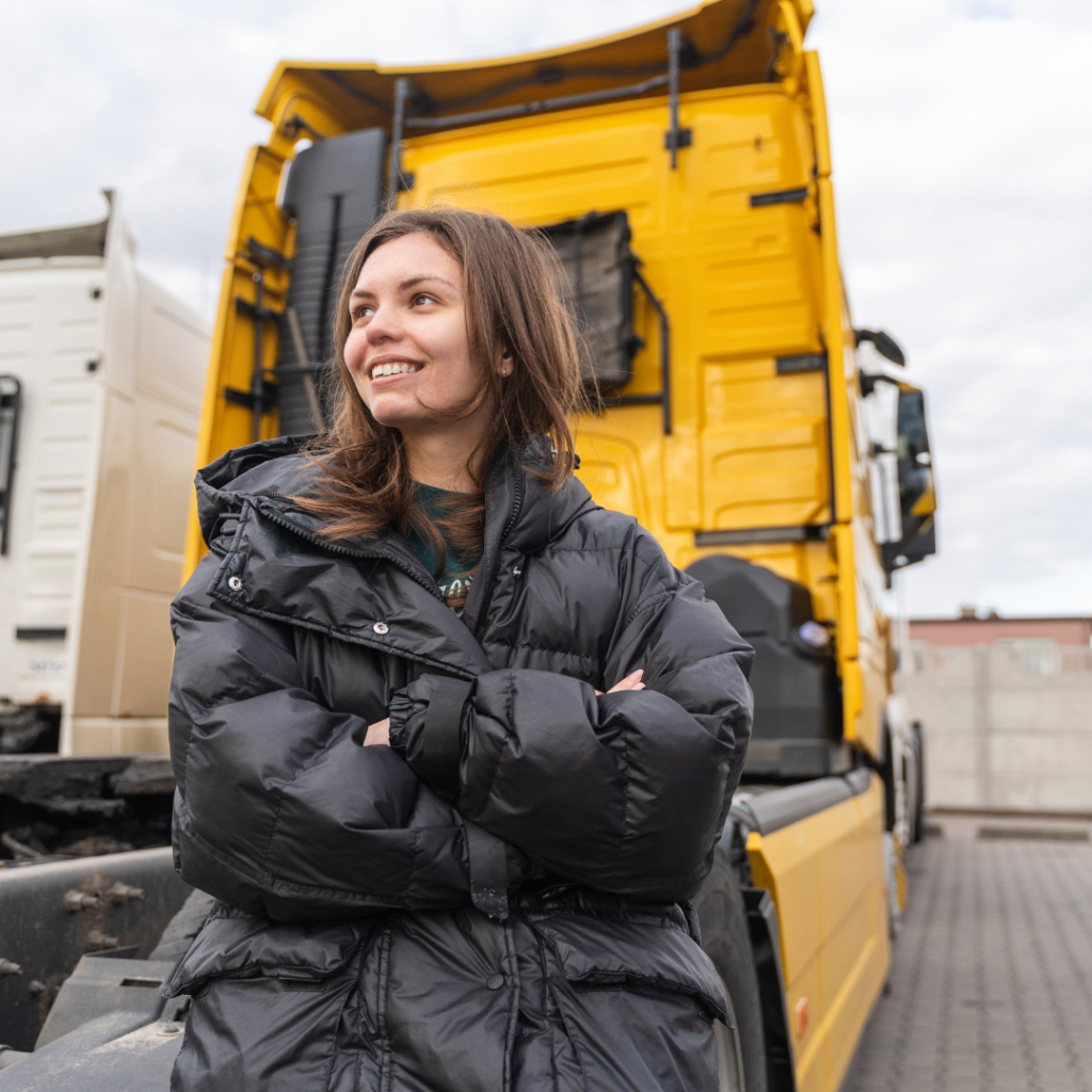 En la imagen se muestra una mujer camionera, con brazos cruzados, recargada en un camión de color amarillo.