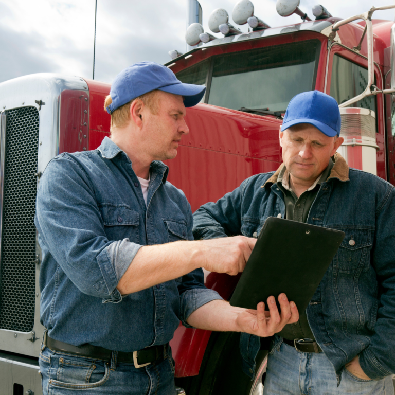 En la imagen se muestran dos hombres frente a un camión, observando papeles en la mano de uno