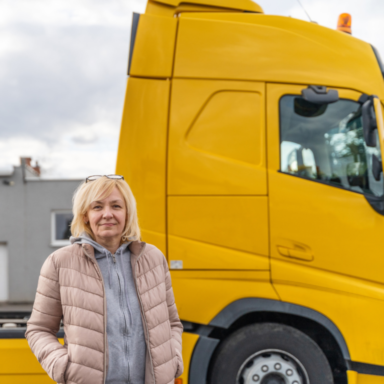 En la imagen se muestra una mujer camionera, parada frente a un camión de color amarillo.